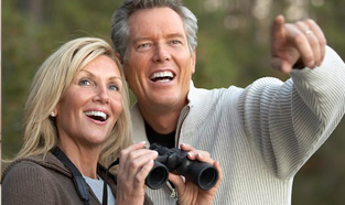 Middle-aged couple outdoors and smiling. The woman is holding a pair of binoculars ready to see what the man is pointing to in the distance.